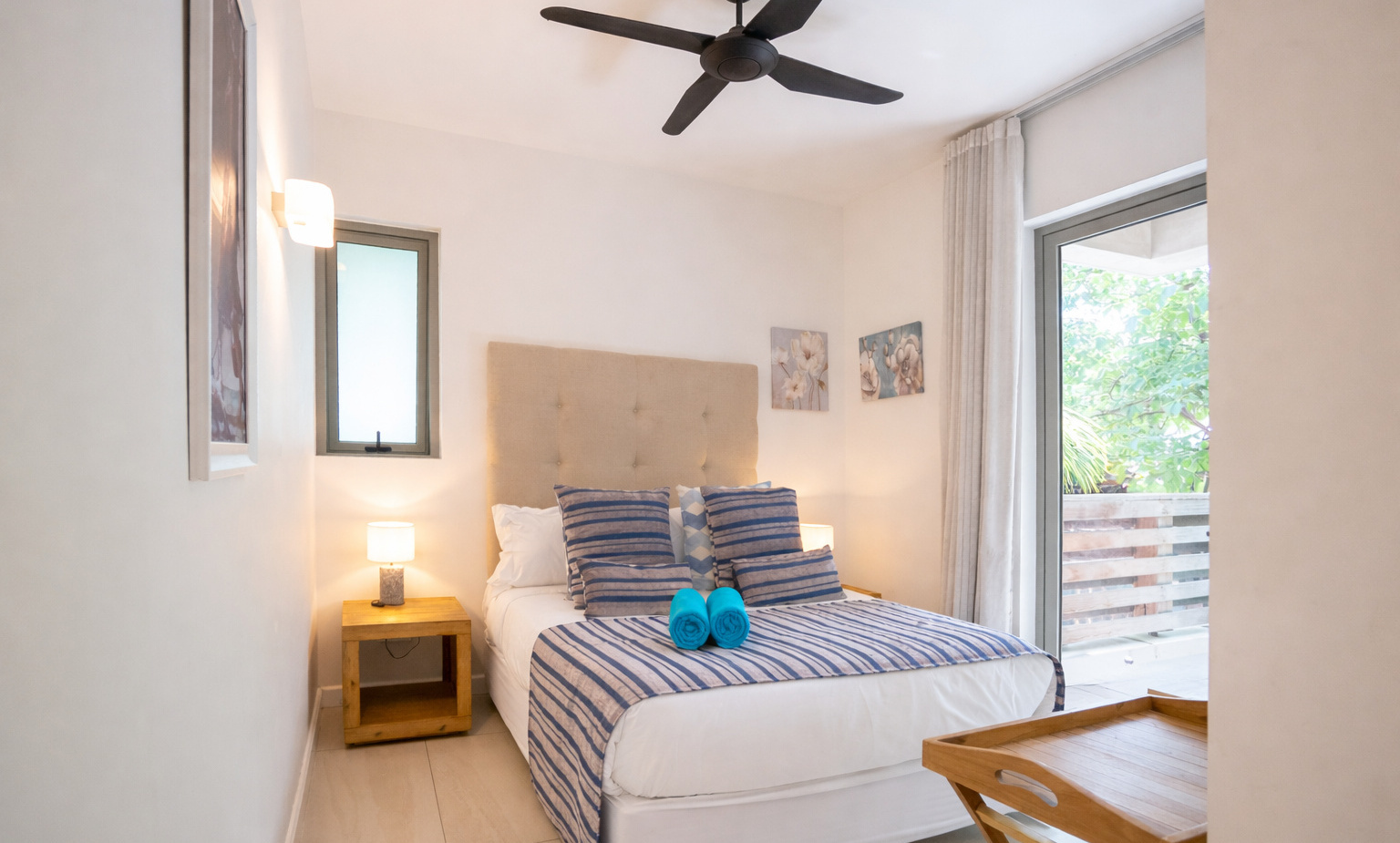 Bright modern double room with tufted headboard, striped blue bedding, rolled turquoise towels, bedside lamps and balcony.