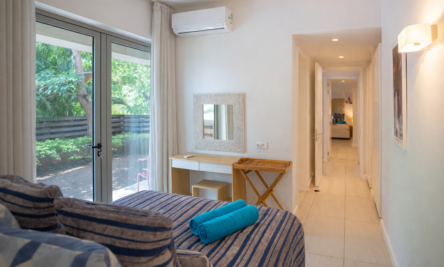 Bright garden-view guest room with sliding glass doors, striped daybed with blue rolled towels, desk, mirror and AC.