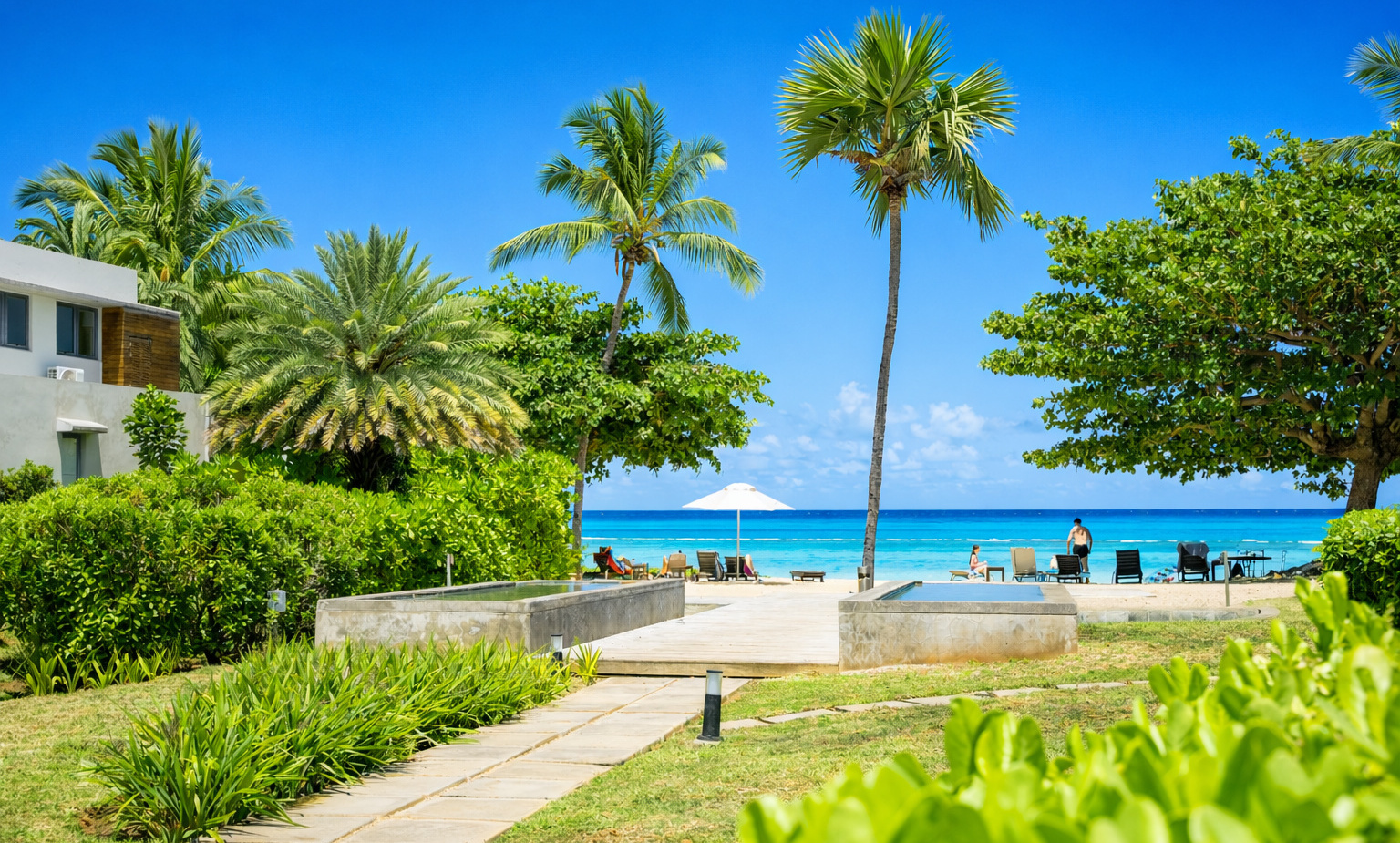 Tropical resort pathway through manicured gardens and palms leading to beachfront loungers, umbrella and turquoise ocean view