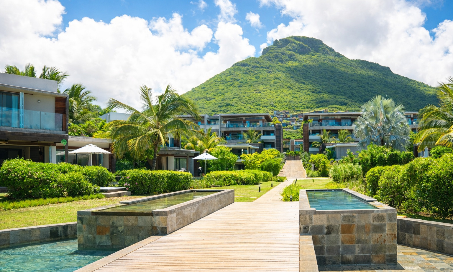 Resort walkway flanked by shallow reflecting pools, tropical gardens and palm-fringed private villas with mountain backdrop and blue sky.