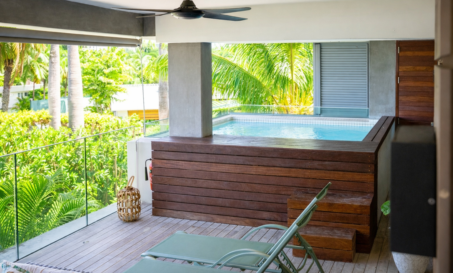 Private balcony with raised wooden plunge pool, two green lounge chairs, ceiling fan and glass railing overlooking tropical palm gardens.