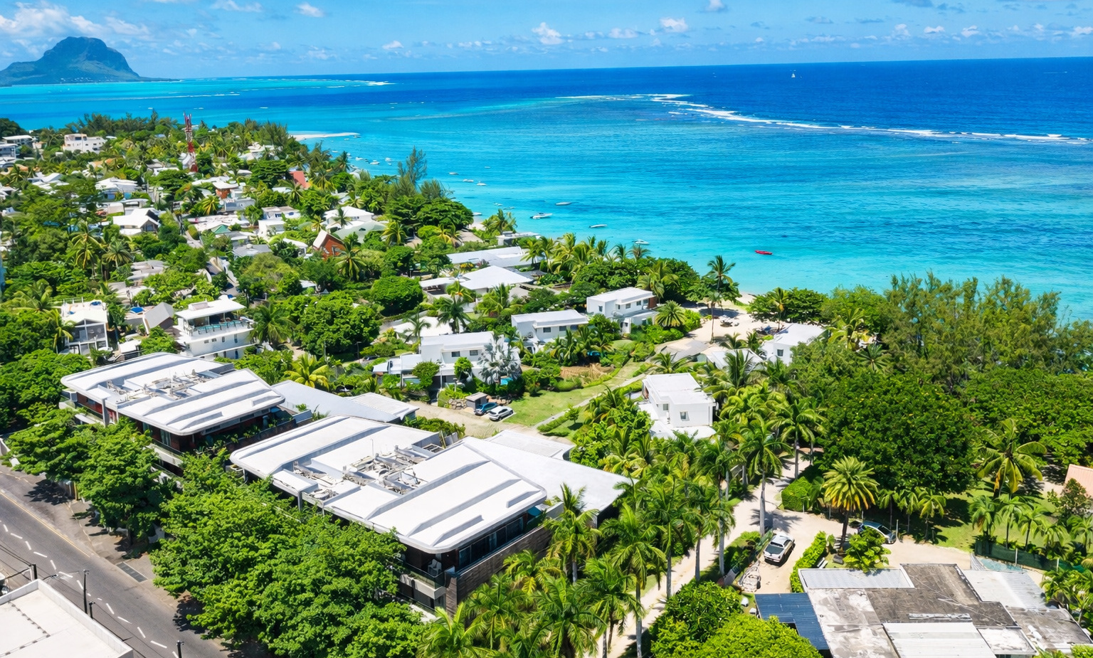 Coastal resort with white villas and palm-lined streets overlooking a turquoise lagoon, boats and distant mountain view