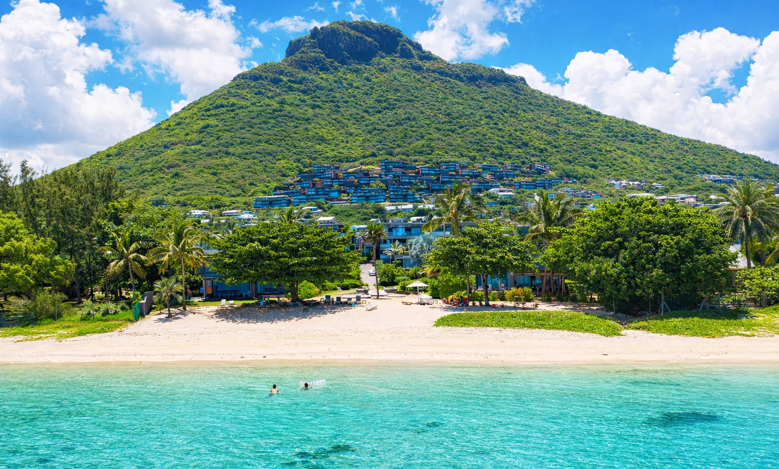 Turquoise lagoon with two guests swimming, sandy beach with loungers and palm trees, resort villas cascading up a lush hillside.