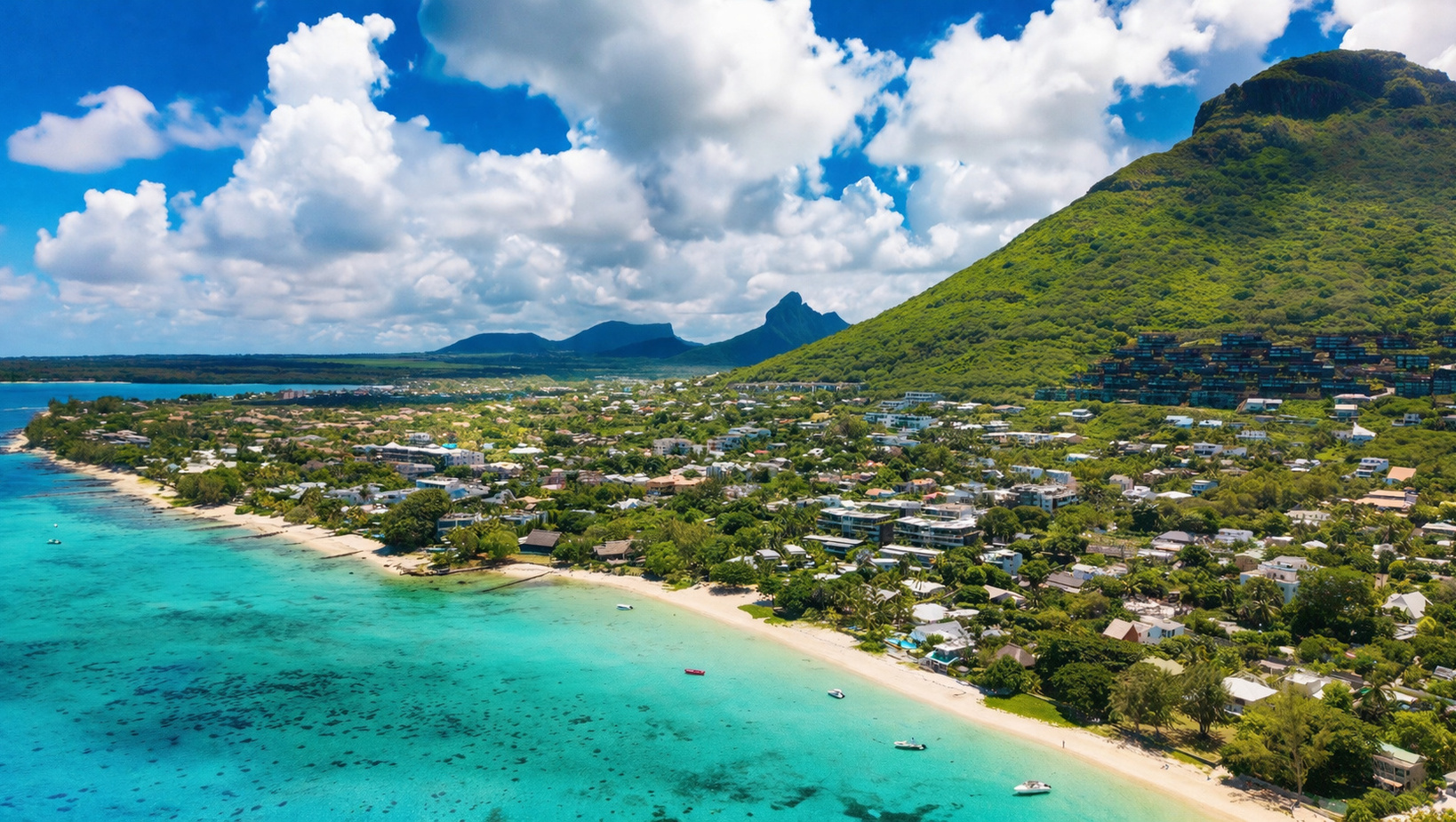 Turquoise lagoon and sandy beach lined with villas and resorts, green mountain rising behind, small boats offshore.