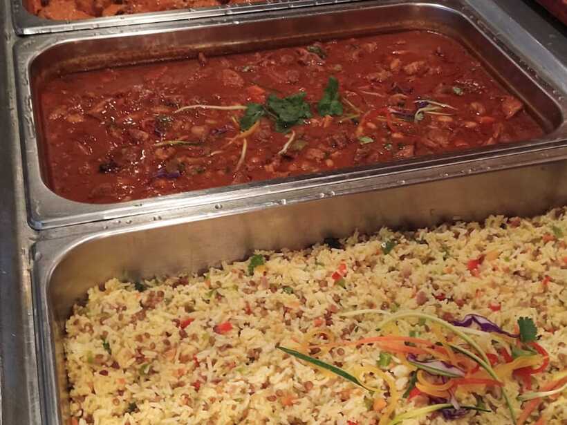 Hotel buffet with rice pilaf garnished with vegetables, two rich tomato curries and vegetable sides in stainless trays.