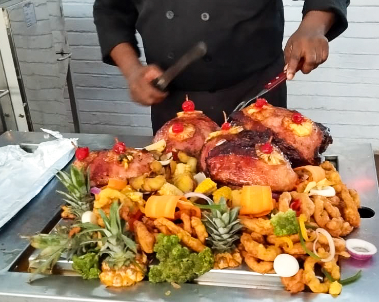 Chef carving glazed pineapple-topped hams on a festive buffet platter with fried sides, pineapple crowns, vegetables and cherries.