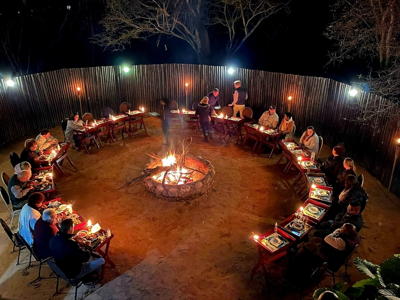 Candlelit outdoor dinner circle around a central stone campfire, guests seated at individual tables inside a wooden fence