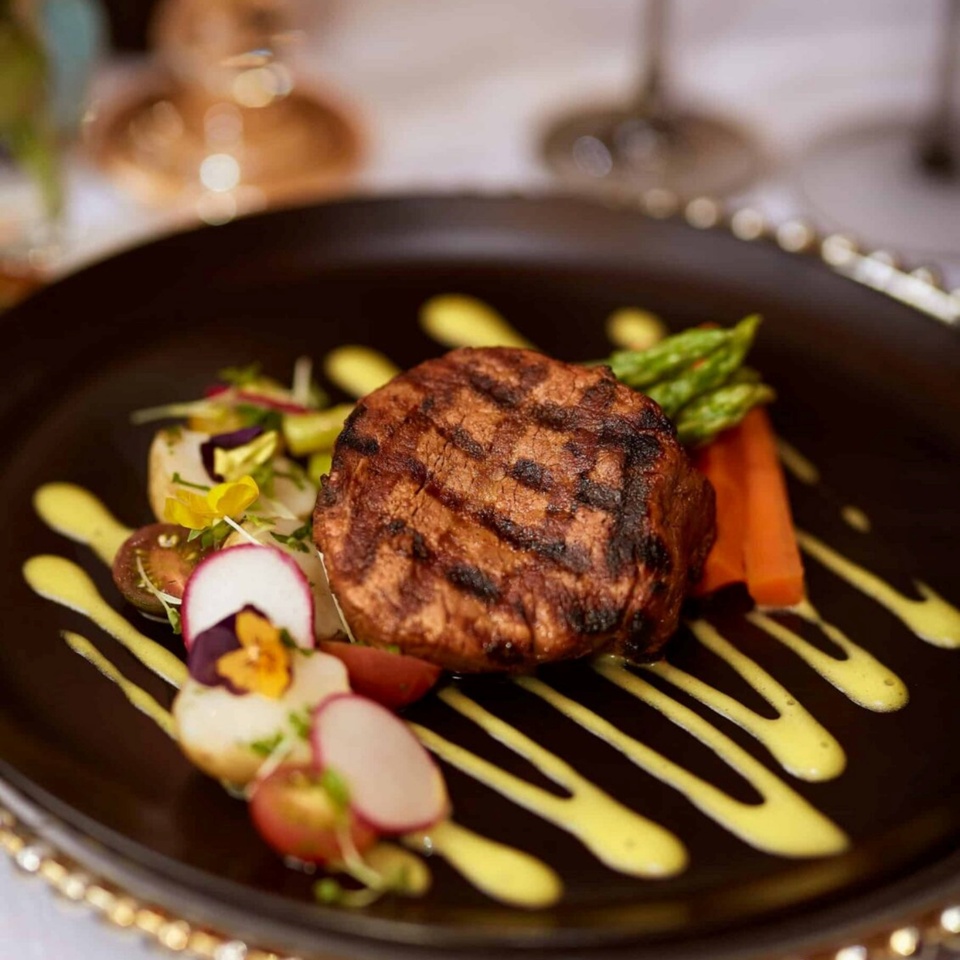 Grilled steak medallion on black plate with yellow sauce, asparagus, carrots and radish slices, elegant fine-dining.