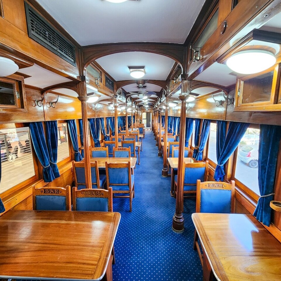 Vintage wooden dining car with polished tables, blue velvet curtains and chairs, aisle seating and soft overhead lighting.