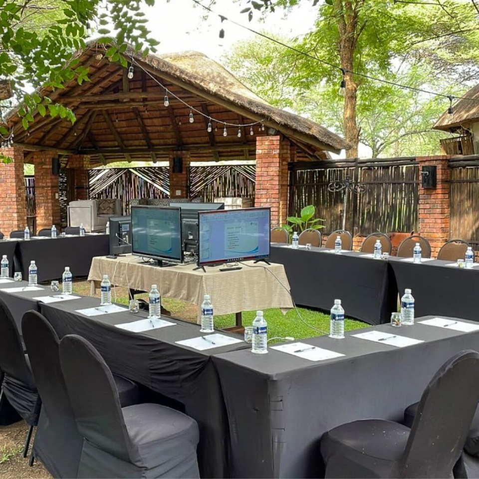 Thatched outdoor meeting space with U-shaped black-draped tables, chairs, bottled water, notepads and central monitors.