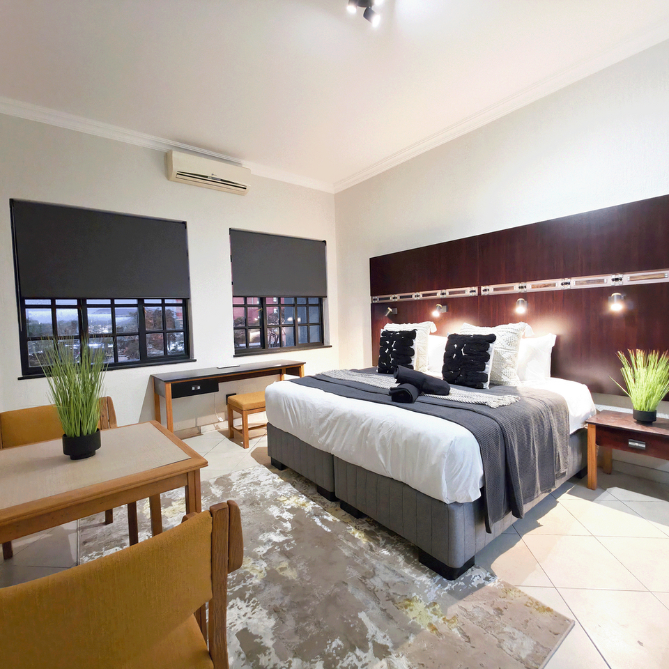 Modern double hotel room with king bed, dark wood headboard, bedside lamps, desk, dining table, city-view windows and AC.