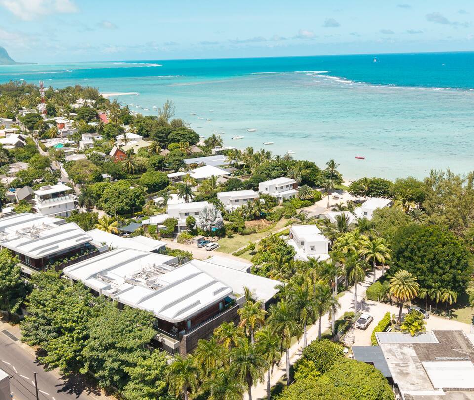 Aerial view of palm-lined beachfront with modern white villas and resort buildings facing turquoise lagoon and anchored boats.