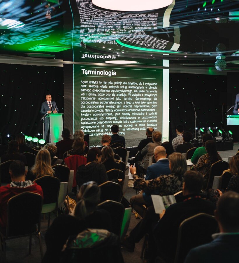 Conference in a modern hall with large screen and speaker presenting agritourism terminology to seated audience.