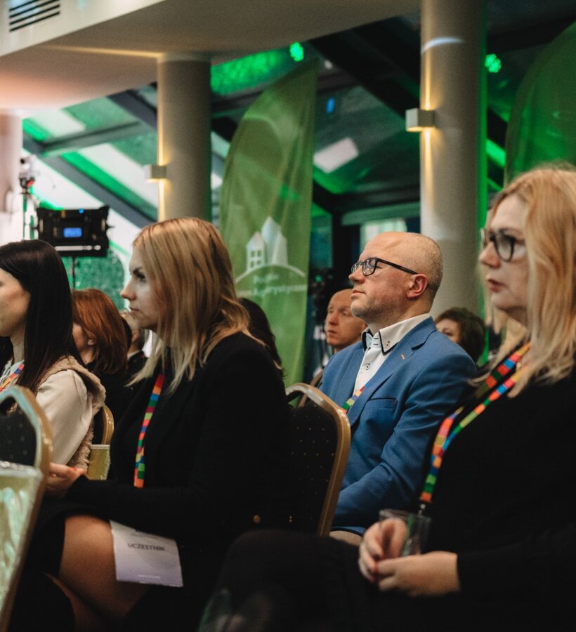 Conference attendees sit attentively in a modern meeting room with green ambient lighting during a presentation.