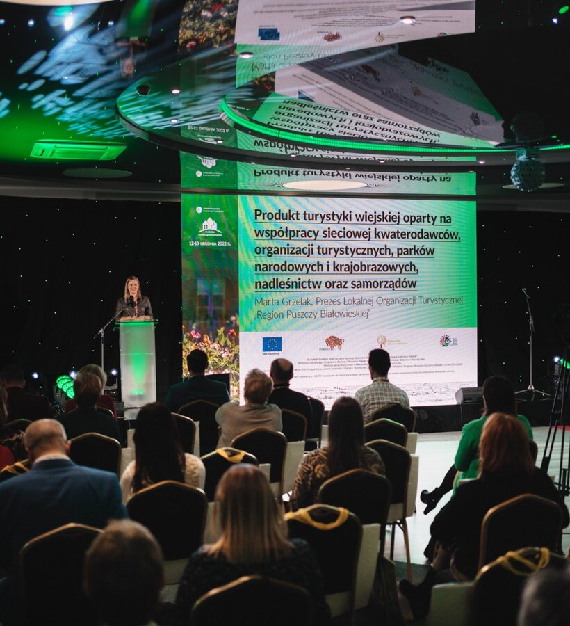 Conference room with stage where a woman is speaking at a tourism industry event, audience seated and attentive