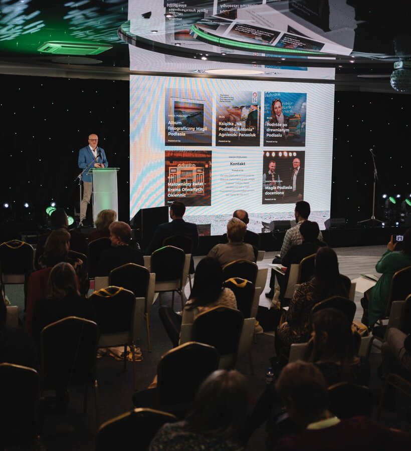 Conference in a modern hall with a speaker at a podium and audience seated, large screens displaying a presentation background.
