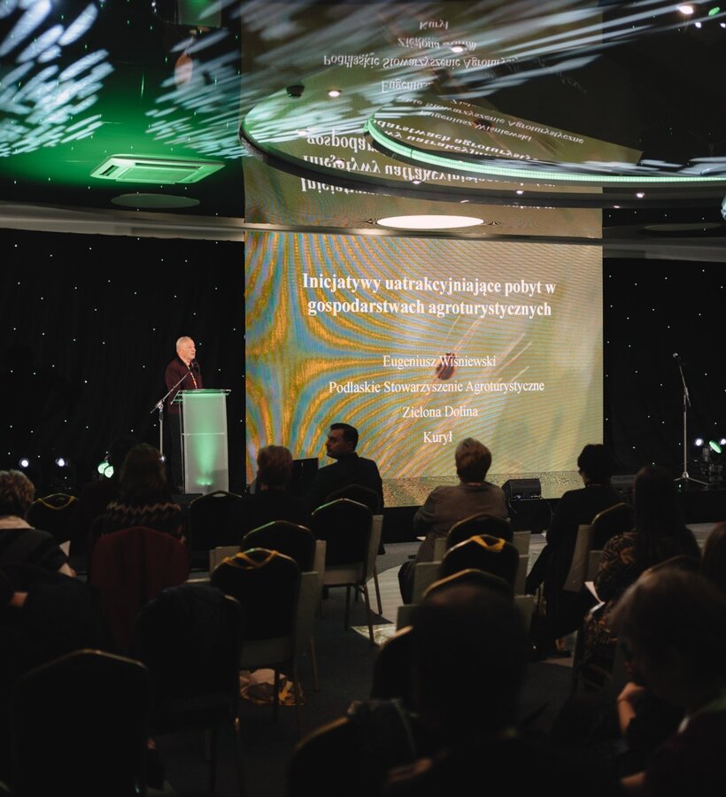 Tourism conference presentation with speaker addressing audience in a dimly lit room featuring large screen and sound system