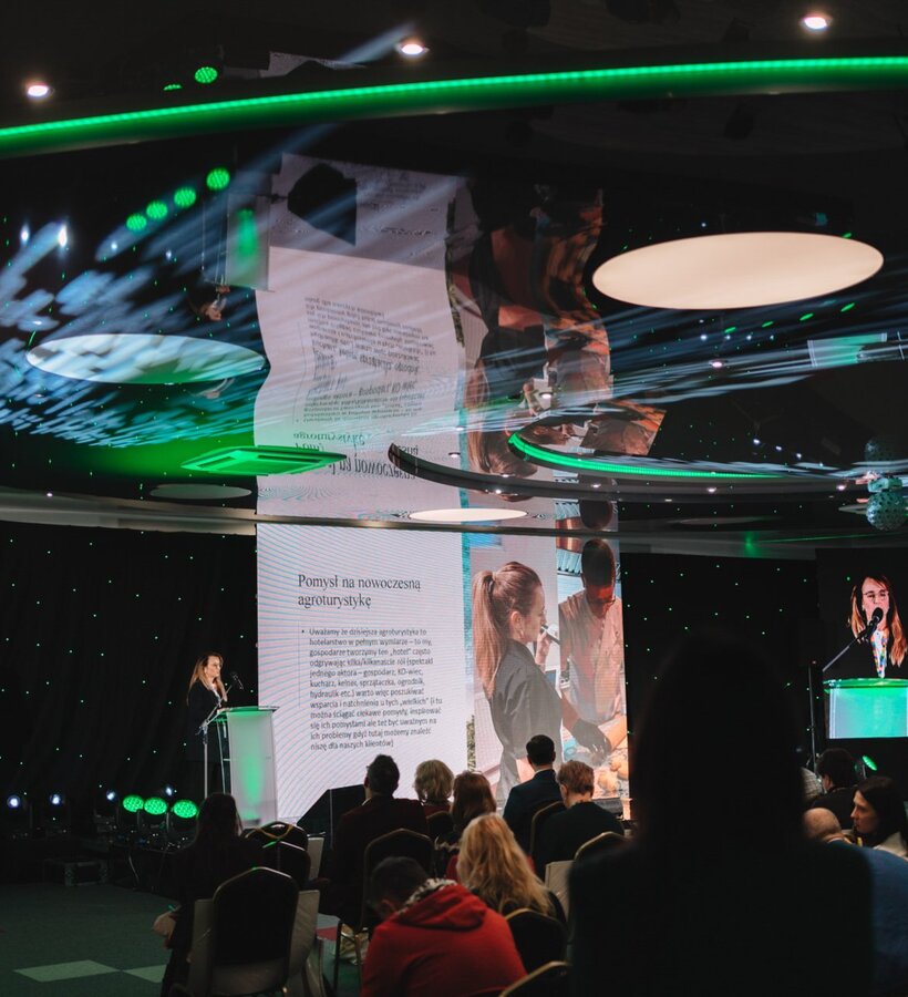 Conference room with woman giving a presentation at illuminated podium, guests seated, modern green LED lighting and projection screens
