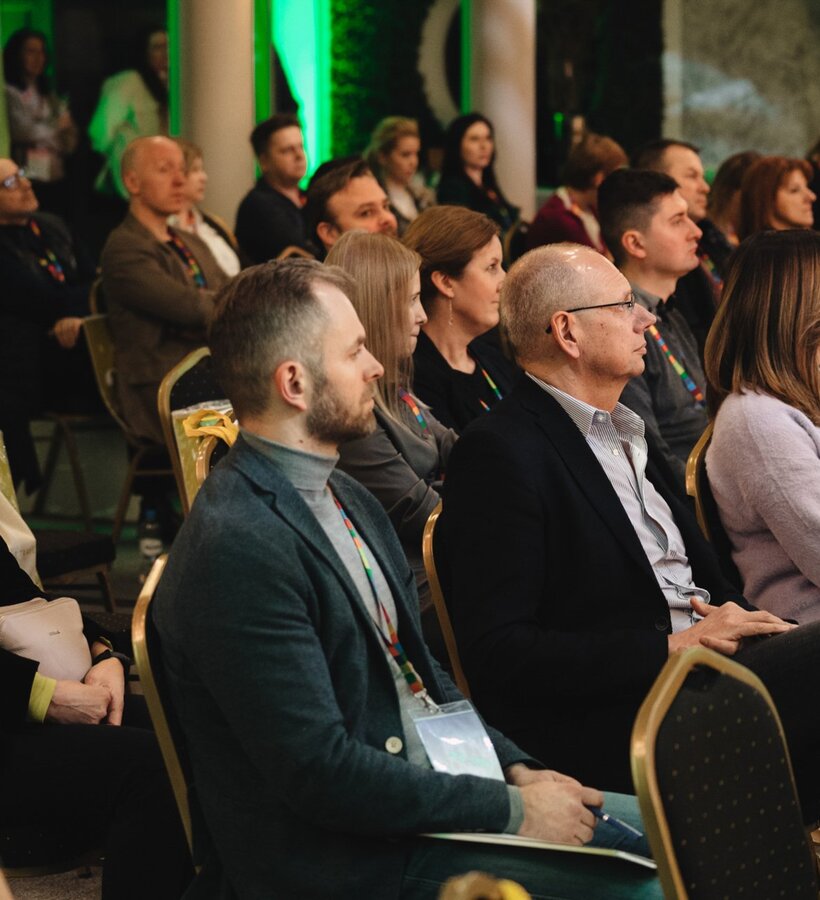 Guests seated attentively at a conference in a warmly lit room with green decorative lighting and formal seating.