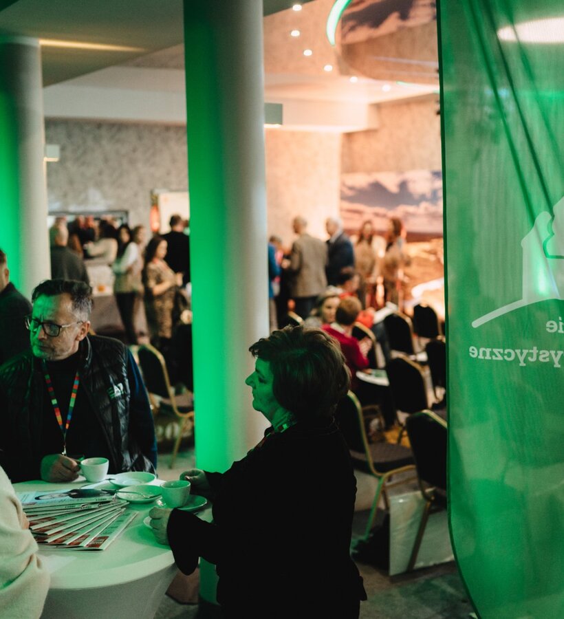 Groups of people chat and drink coffee at a high table during a conference in a spacious room with green lighting.