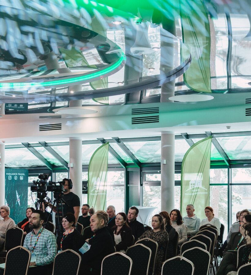 Modern conference room with large windows, reflective ceiling, green flags, and guests attending an event.