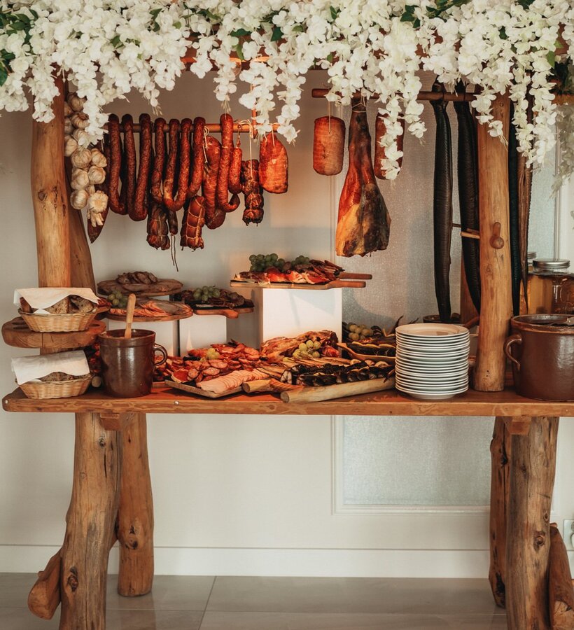 Rustic buffet with dried meats, sausages, bread, and grapes, decorated with white flowers hanging over a wooden table.