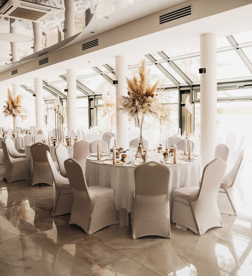 Elegant banquet hall with large windows, white table linens, and decorative pampas grass centerpieces on each table