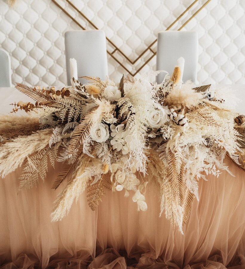 Elegant wedding table decoration with beige and white dried flowers and candles, set against a quilted wall panel backdrop.