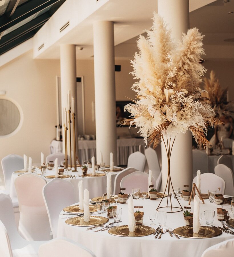 Elegant banquet hall with white tablecloths, gold chargers, and dried flower centerpieces in a bright, modern setting