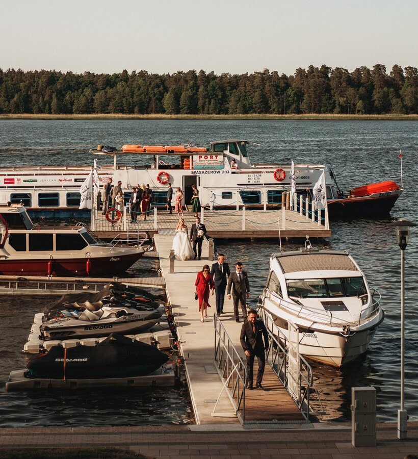 Guests in formal attire disembarking from a passenger boat onto a lakeside dock surrounded by forest