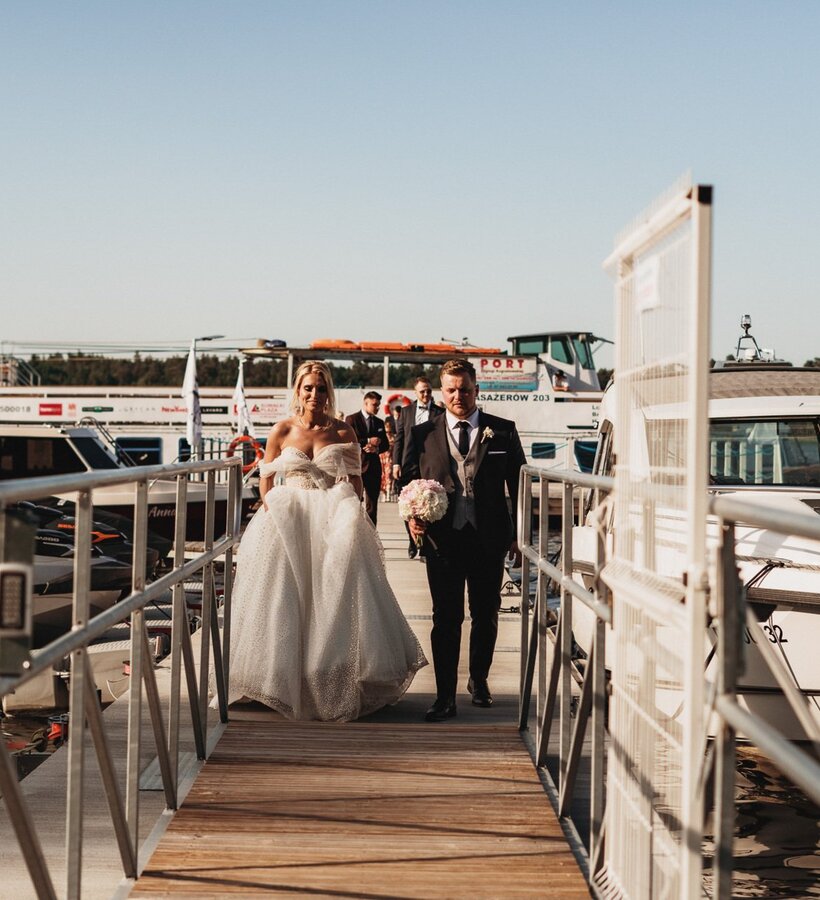 Bride and groom walk along a wooden pier in a marina surrounded by luxury boats on a sunny day.