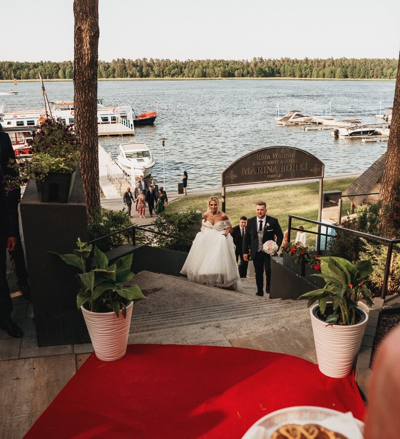 Bride and groom ascending stairs to lakeside hotel, guests dressed elegantly, marina with boats visible in background