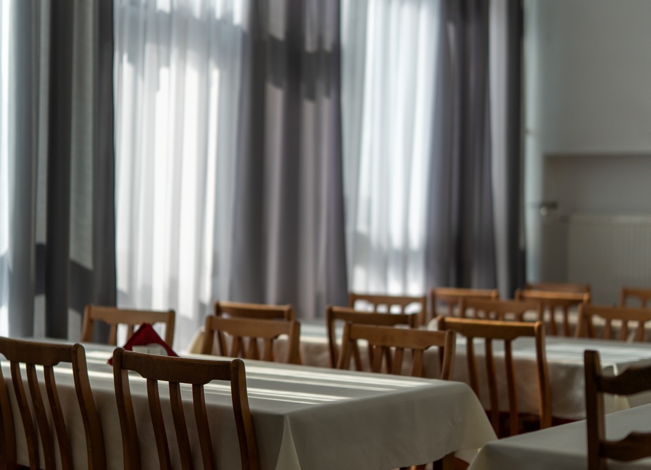 Jadalnia hotelowa z długimi stolikami w białych obrusach, drewnianymi krzesłami i dużymi oknami z półprzejrzystymi zasłonami.