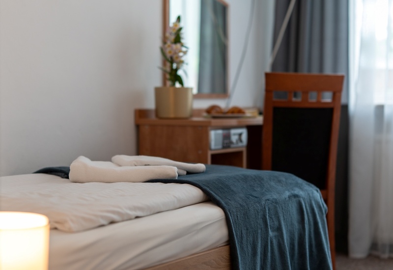Pokój hotelowy z pojedynczym łóżkiem, granatową narzutą i złożonymi ręcznikami, lampką nocną, biurkiem z lustrem, storczykiem i telewizorem.