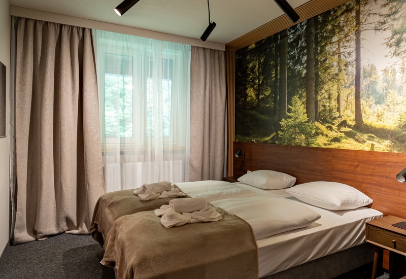 Przytulny pokój hotelowy z dwoma łóżkami, drewnianym zagłówkiem i fototapetą lasu, stolikami nocnymi, lampkami przy łóżku i telewizorem