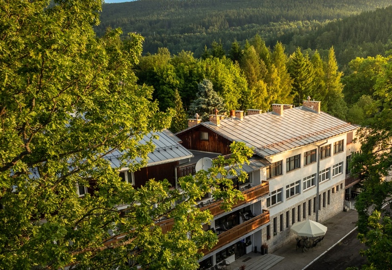 Górski hotel z drewnianymi balkonami i kamienną kondygnacją, taras z parasolem, widok na zielony las i pasmo wzgórz.