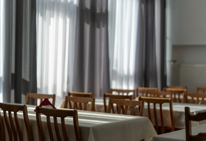 Jadalnia hotelowa z długimi stolikami w białych obrusach, drewnianymi krzesłami i dużymi oknami z półprzejrzystymi zasłonami.