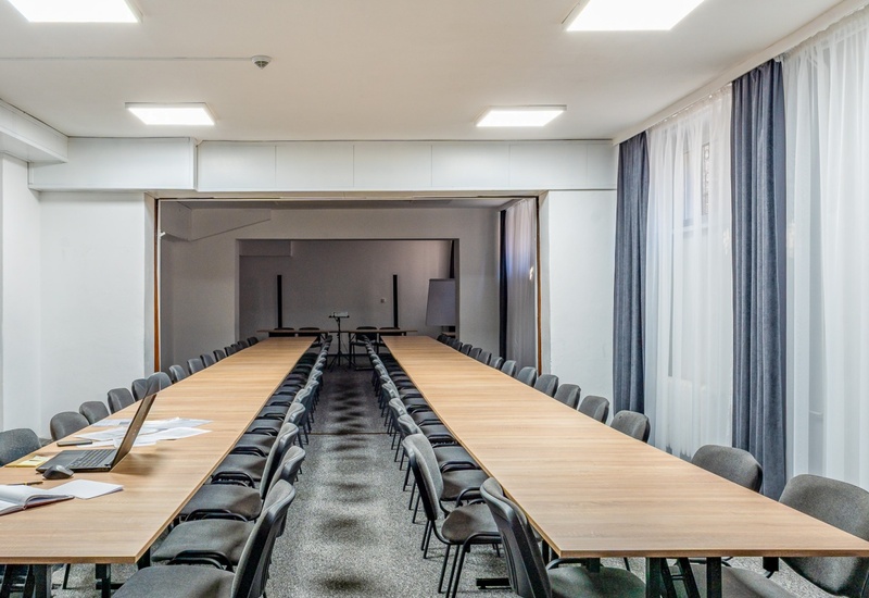 Przestronna sala konferencyjna z długimi drewnianymi stołami, rzędami szarych krzeseł, laptopem i zasłonami.
