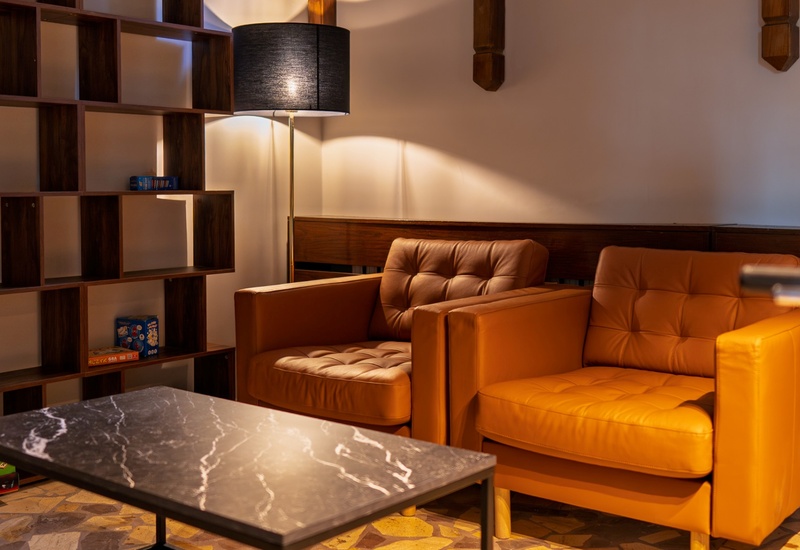 Przytulny hotelowy salonik z dwoma skórzanymi fotelami, marmurowym stolikiem, stojącą lampą i półką z grami planszowymi.