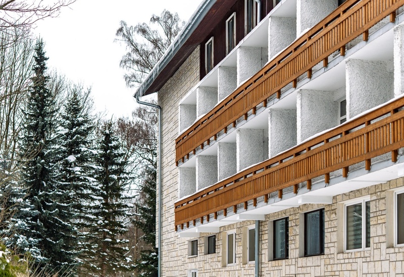 Zimowy hotel z rzędem drewnianych balkonów, kamienną elewacją, śnieżnym podjazdem i sosnami w tle.