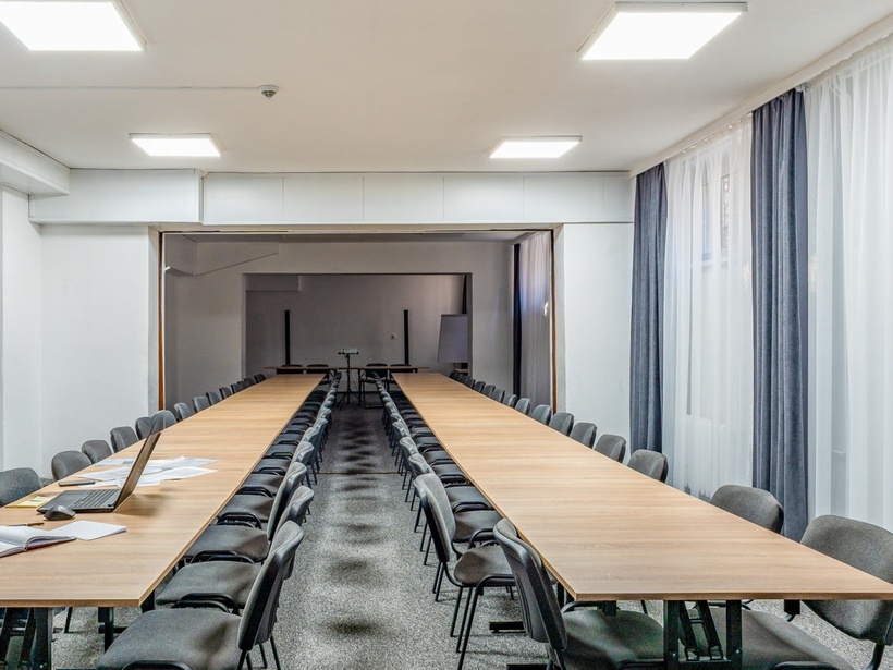 Przestronna sala konferencyjna z długimi drewnianymi stołami, rzędami szarych krzeseł, laptopem i zasłonami.