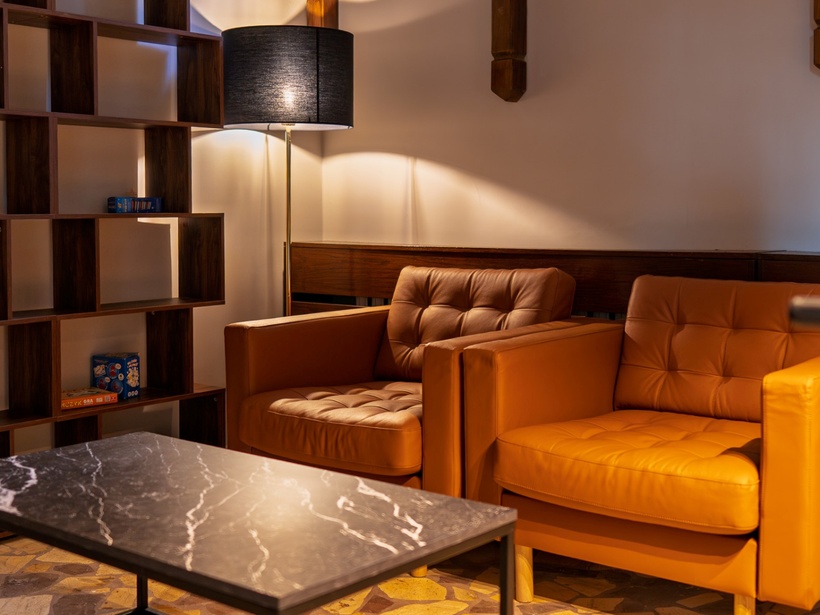 Przytulny hotelowy salonik z dwoma skórzanymi fotelami, marmurowym stolikiem, stojącą lampą i półką z grami planszowymi.