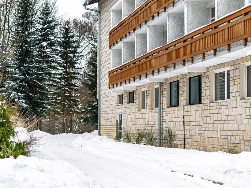 Zimowy hotel z rzędem drewnianych balkonów, kamienną elewacją, śnieżnym podjazdem i sosnami w tle.