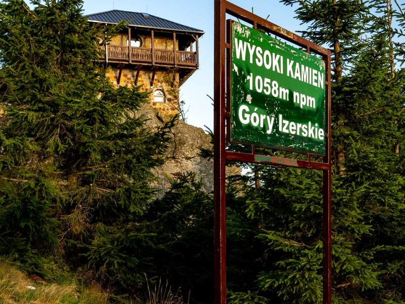 Kamienna wieża‑punkt widokowy z drewnianym balkonem na skale wśród świerków; tablica 'Wysoki Kamień 1058 m n.p.m., Góry Izerskie'.