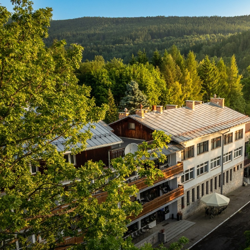 Górski hotel z drewnianymi balkonami i kamienną kondygnacją, taras z parasolem, widok na zielony las i pasmo wzgórz.