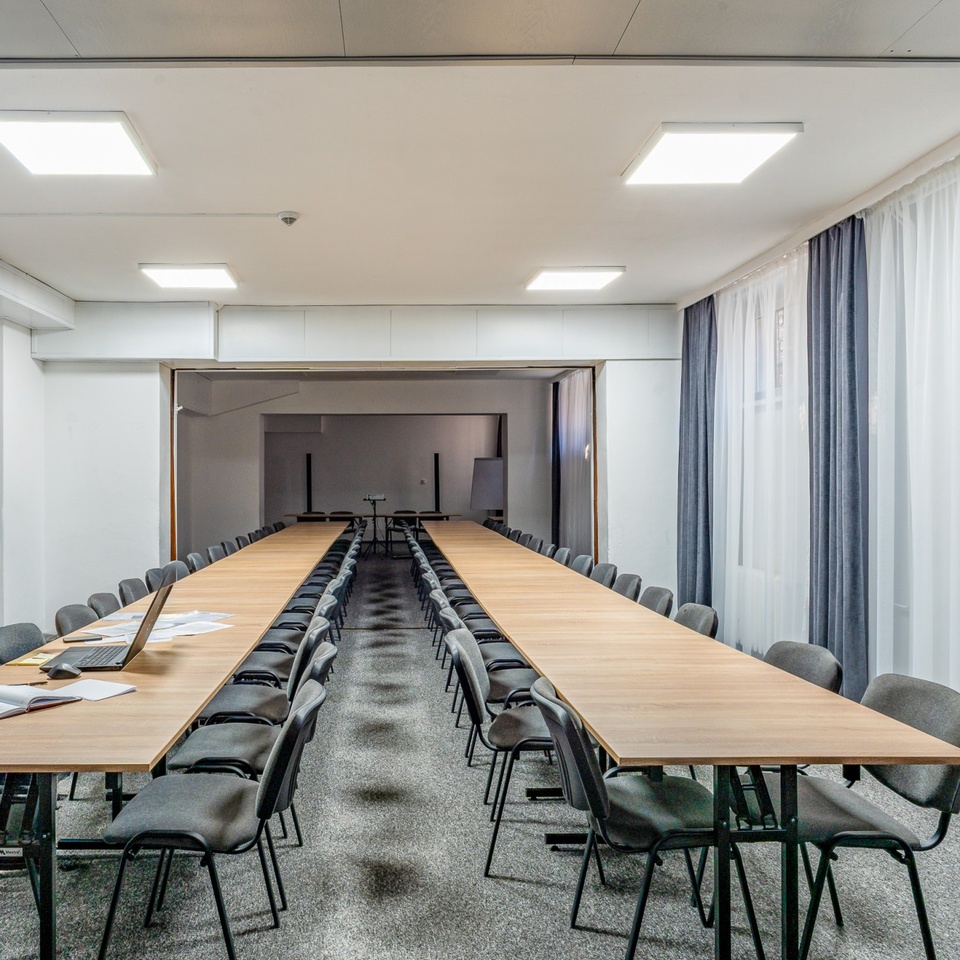 Przestronna sala konferencyjna z długimi drewnianymi stołami, rzędami szarych krzeseł, laptopem i zasłonami.