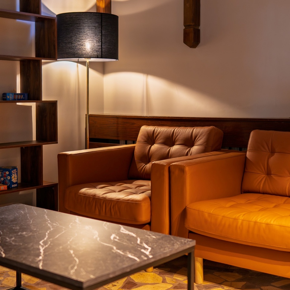 Przytulny hotelowy salonik z dwoma skórzanymi fotelami, marmurowym stolikiem, stojącą lampą i półką z grami planszowymi.