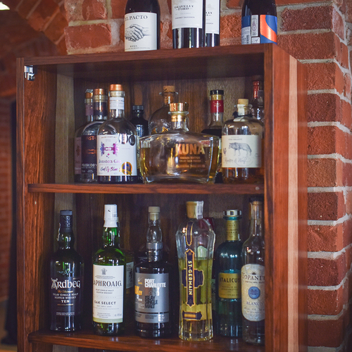 Holzregal der Bar vor freigelegtem Backstein mit Wein- und Spirituosenflaschen sowie gestapelten Gläsern, gemütliches Hotelbar-Ambiente