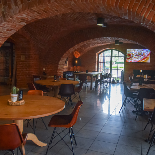 Ziegelgewölbe-Restaurant mit runden Holztischen, Lederstühlen, Bogenfenster zum Garten, warmem Licht und Wand-TV mit Pizzabild.