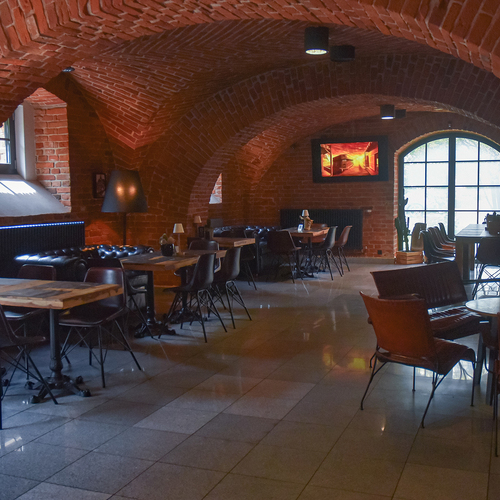 Gewölbte Ziegel-Lounge mit Rundbögen, Ledersofas, Holztischen und warmer Beleuchtung am großen Rundbogenfenster
